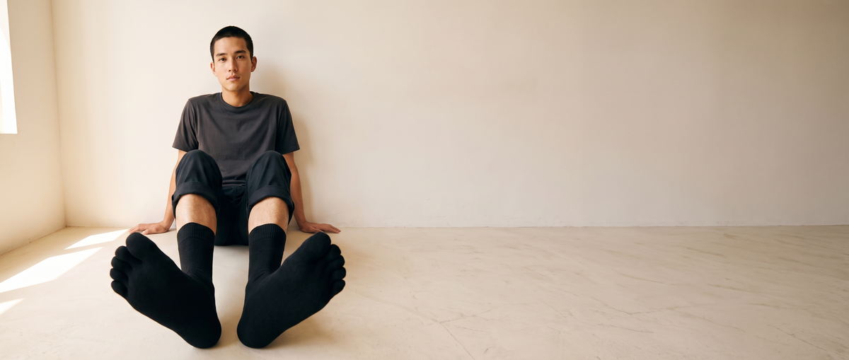 A man seated on a pale floor with his legs extended toward the camera, wearing ROMATOKI merino five-toe socks in Coal